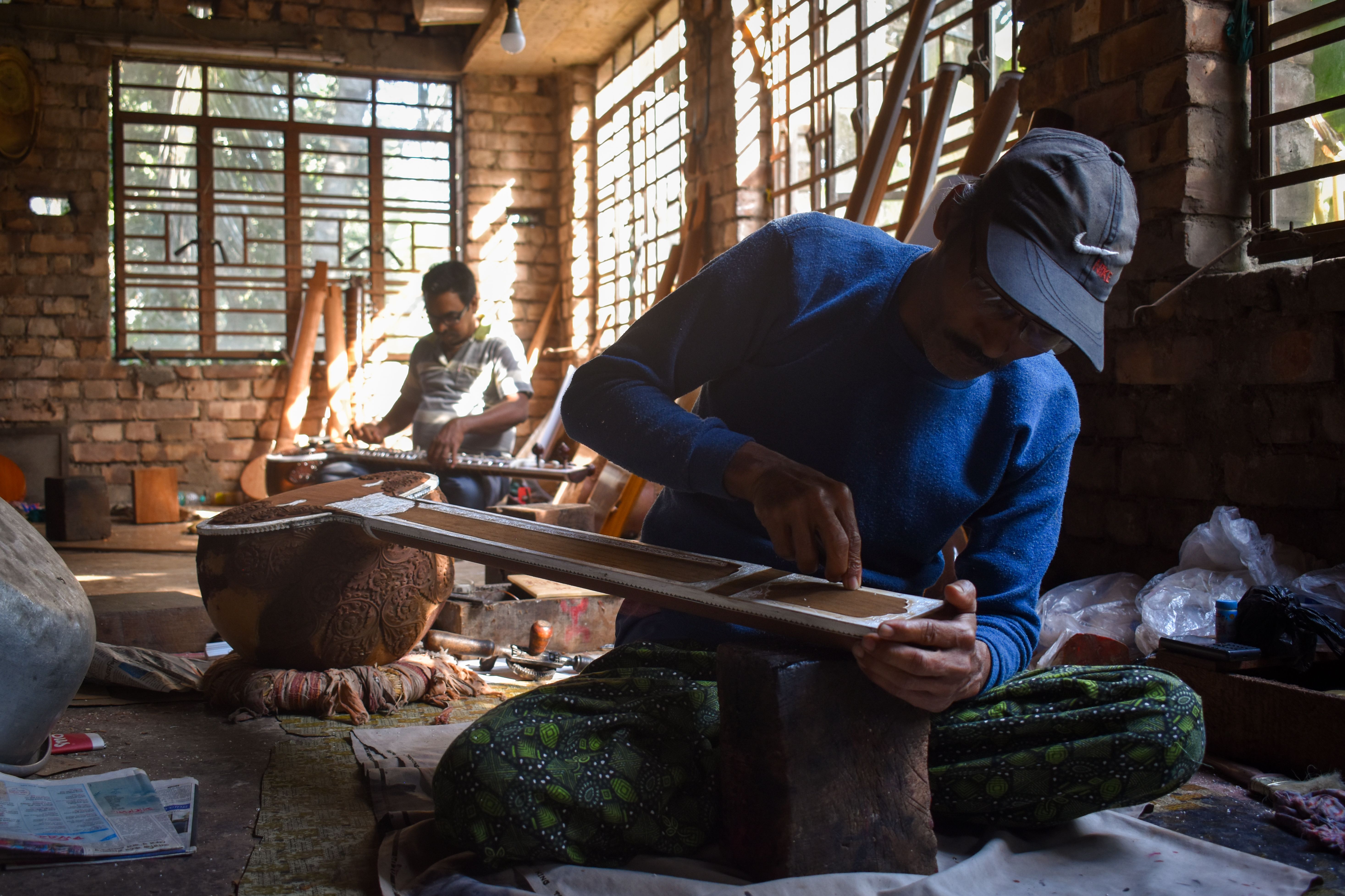 An Indian master luthier in his Kolkata workshop applying the final polish to a sitar neck, surrounded by tools and half-finished instrument bodies in warm workshop light