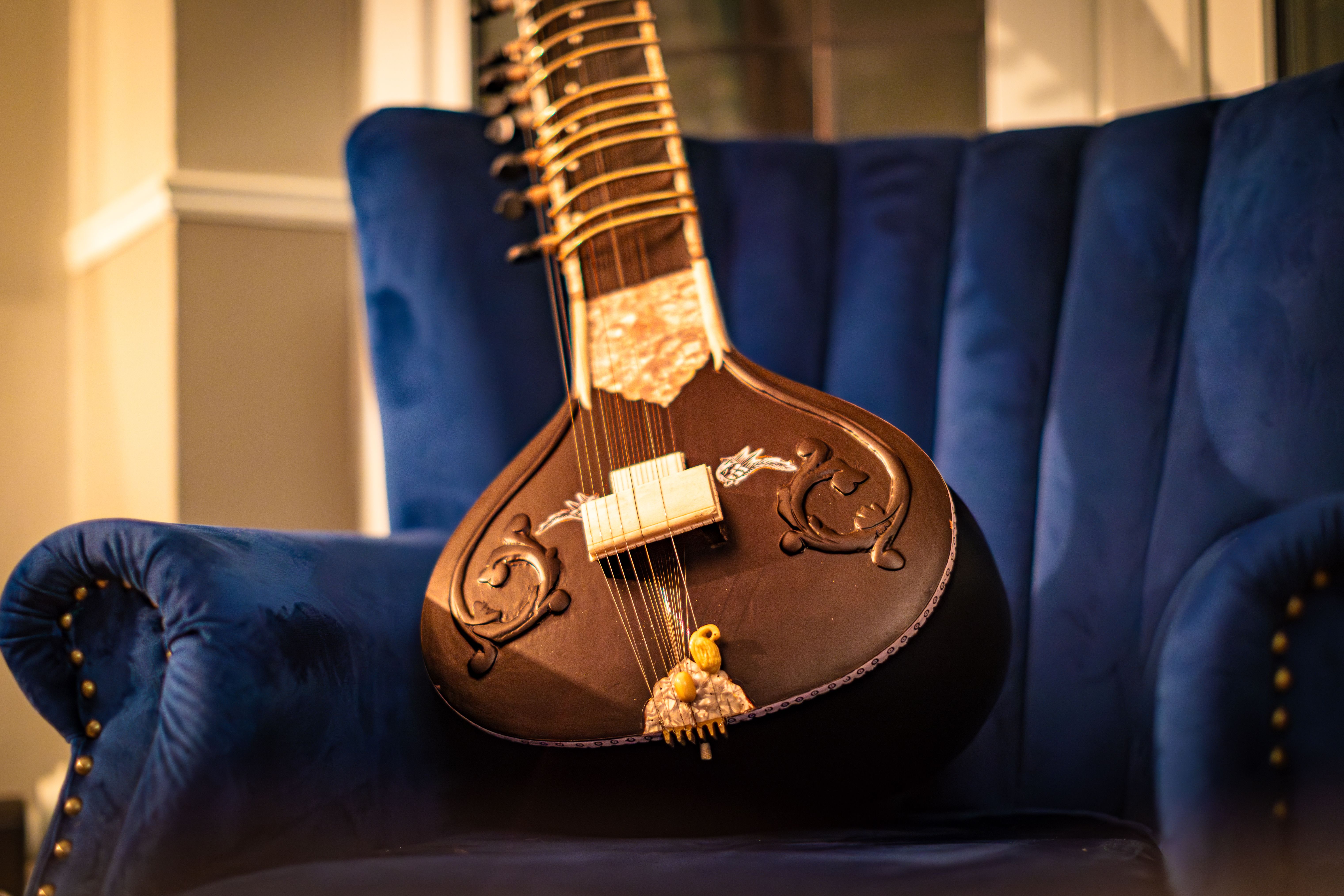 A beautifully crafted sitar resting against a wall in warm amber studio light, its teak neck and gourd body showing the detailed hand-carved decorative work of a Kolkata master luthier